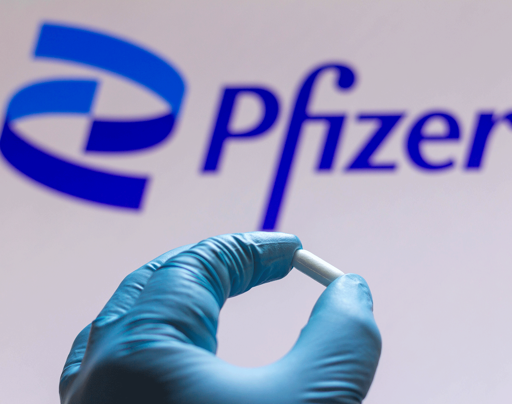 Gloved hand holding a white pill in front of the Pfizer logo.